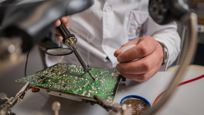 Modyfikacja Radeona 9500 lutownicą Technik w białym fartuchu lutuje zieloną płytkę drukowaną